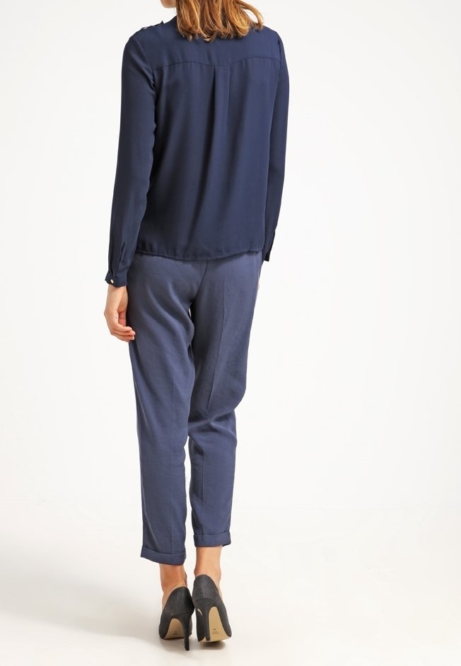 Blusa blu navy con maniche lunghe e schiena plissettata, abbinata a pantaloni navy a gamba stretta. L'outfit è completato da scarpe con tacco alto nere.