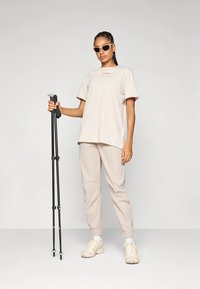 Beige bomulls t-skjorte med liten rød logo, beige avsmalnende bukser og beige joggesko. Modellen holder svarte vandrestaver og har på seg solbriller.