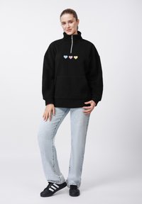 Μαύρο φλις πουλόβερ με φερμουάρ στη μέση, διακοσμημένο με τρία απλικέ σε παστέλ αποχρώσεις καρδιές. Συνδυάζεται με ανοιχτό μπλε τζιν και μαύρα αθλητικά παπούτσια.