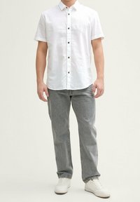 Weißes Kurzarm-Hemd mit Knöpfen aus strukturiertem Stoff, kombiniert mit grauen, geraden Jeans und weißen Sneakers. Einfaches Design mit braunen Knöpfen.