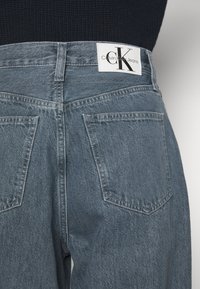 Vaaleansiniset farkut, joissa on korkea vyötärö, kaksi takataskua ja valkoinen etiketti, jossa on "Calvin Klein Jeans" -logo näkyvissä.