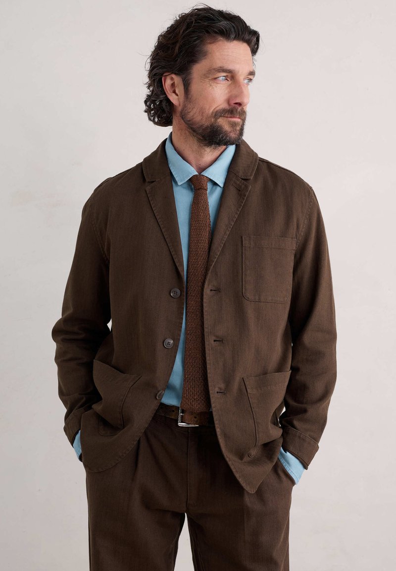 Brun bomuldsblazer med notched krave, frontlomme, parret med en lyseblå skjorte og en struktureret brun slips.