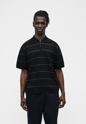 Muž s krátkymi dredmi, oblečený v čiernej polokošeli s krátkym rukávom a tenkými horizontálnymi pruhmi, stojaci pred jednoduchým svetlým pozadím.