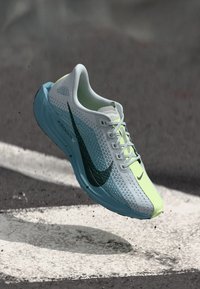 Gaiši zils un neona zaļš Nike skriešanas apavs ar melnu Swoosh logo, karājoties virs asfalta ar baltu krāsotu līniju.