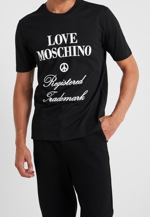 Mies pukeutunut mustaan t-paitaan, jossa valkoinen teksti "LOVE MOSCHINO" ja "Registered Trademark" sekä rauhansymboli, yhdistettynä mustiin housuihin.