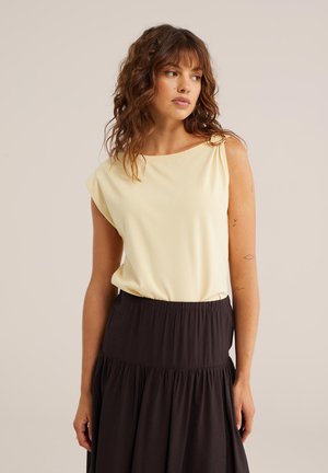 Femeie cu păr castaniu ondulat, purtând un top crem fără mâneci, băgat într-o fustă neagră cu talie înaltă, uitându-se ușor spre stânga.