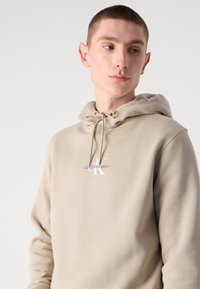 Bežna kapuca iz mehkega materiala, ki ima nastavljivo kapuco in logo z napisom "Calvin Klein Jeans" v kontrastnih barvah.