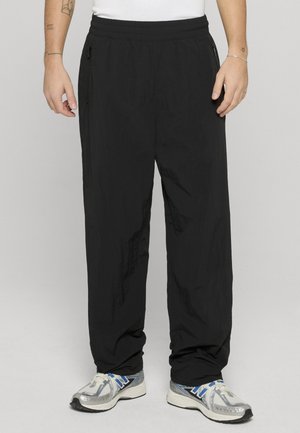 Persona con pantalones negros de corte holgado con bolsillos con cremallera y zapatillas de correr plateadas con detalles azulados, de pie frente a un fondo liso.