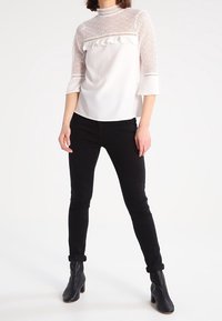 Blusa bianca con dettagli in pizzo e accentuazione arricciata sul petto, abbinata a pantaloni neri slim-fit e stivaletti alla caviglia.