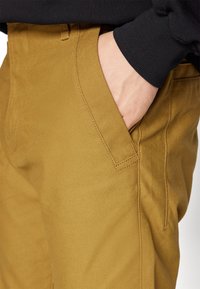 Bruna byxor med en slät yta och rak skärning. Funktioner inkluderar sidofickor och synliga sömnadsdetaljer. En svart ärm är delvis synlig.