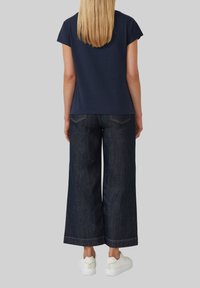 Marineblauwe korte mouwen top gecombineerd met wijd-uitlopende donkere denim jeans. Witte sneakers maken de outfit af. Achteraanzicht met zichtbare stikdetails.