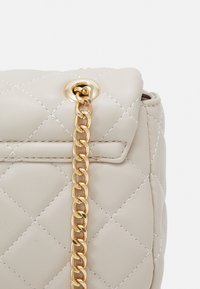 Bolso acolchado beige hecho de cuero sintético, con una correa de cadena dorada y un cierre de solapa con un acento metálico circular.