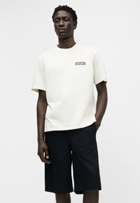 Άνδρας που φοράει λευκό t-shirt Dickies και μαύρο σορτς μήκους μέχρι το γόνατο, στέκεται με το ένα χέρι στην τσέπη μπροστά σε απλό φόντο.