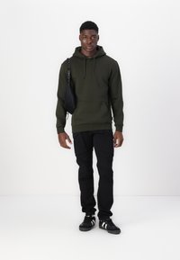 Olive green hooded sweatshirt with front pocket, paired with black cargo pants and black shoes featuring white stripes and adjustable strap bag.