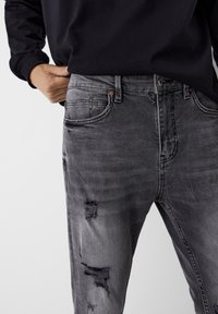 Gray denim jeans with distressed areas, five pockets, button closure, and contrasting stitching. Worn with a black long-sleeve top.