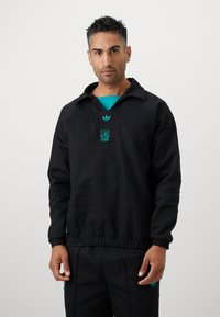 Sort pullover med en afslappet pasform, der har krave og elastisk talje. Smykket med turkis Adidas-logo og Liverpool-våbenskjold.