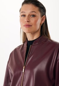 Giubbotto bomber in pelle borgogna con una finitura lucida e una zip dorata. Presenta un colletto rialzato e un design con vestibilità rilassata.