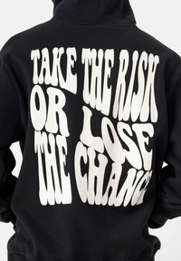 Sudadera negra con un gran texto blanco en la parte trasera, que dice "TOMA EL RIESGO O PIERDE LA OPORTUNIDAD" en una fuente gruesa y ondulada.