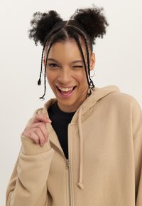 Young woman with braided hair and two afro puffs winks and smiles, wearing hoop earrings, a black top, and a beige zip-up hoodie.