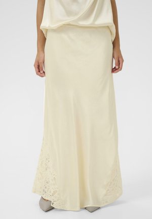 Kobieta ubrana w długą, kremową spódnicę satynową z koronkowymi zdobieniami przy krawędzi, w połączeniu z szarymi, spiczastymi butami na białym tle.