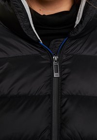 Sort pufferjakke med lynlås foran, med blå accenter på lynlåsen og en grå inderforing. Glat, quiltet tekstur.