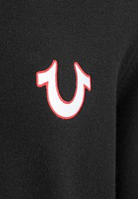 Schwarzer Stoff mit einer strukturierten Oberfläche, der ein markantes weißes und rotes gesticktes Logo in Form einer doppelten Kurve aufweist.