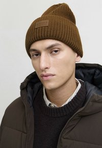 Un jeune homme portant un bonnet en tricot marron, un pull sombre par-dessus une chemise à col, et un blouson matelassé marron avec fermeture éclair, sur un fond uni.