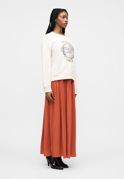 Γυναίκα με μακριά πλεξούδες που φοράει λευκό φούτερ με σχέδιο, μακρύ φούστα σκουρόχρωμης απόχρωσης και μαύρα μποτάκια με τακούνι, όρθια σε προφίλ.