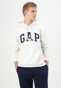Sweatshirt crème avec le logo "GAP" bleu marine, capuche avec cordon et poche avant. Associé à un pantalon bleu marine, avec une coupe décontractée.