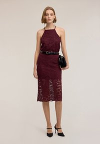 Viininpunainen pitsimekko, jossa on halterneck, istuva bodi ja polvenpituinen hame. Varustettu mustalla vyöllä ja pikkulaukulla. Kengät ovat mustat, joissa on remmit.