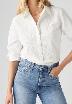 Kvinde iført en hvid langærmet bluse med broderede detaljer foran og lyseblå højtaljede denimjeans, den ene hånd i lommen.