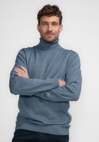 Blauwe coltrui van zacht breimateriaal met geribde manchetten en onderrand, gedragen met gekruiste armen voor een effen achtergrond.