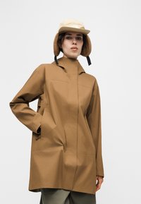 Persona valkāš brūnganu ūdensizturīgu jaku un bēša zimaiņu cepuri ar ausu aizsegu vienkārša gaiši pelēka fona priekšā.