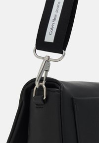 Czarna skórzana torba o teksturowanym wykończeniu; wyposażona w metalowy klips i czarny pasek z napisem "Calvin Klein Jeans". Ostro zarysowane kąty i elegancki design.