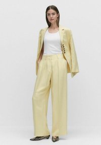 Blazer tailleur jaune avec un pantalon large assorti, complété par un débardeur blanc et des chaussures en serpent, mettant en avant une texture douce et lisse.