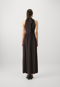 Robe halter noire longue avec un dos noué et une taille froncée, en tissu lisse. Paire de sandales noires à talons carrés visibles.