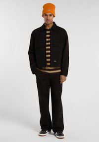 Mladý muž stojí sebevědomě v černé bundě přes pruhovanou košili, černých kalhotách a teniskách, na hlavě má oranžovou čepici.