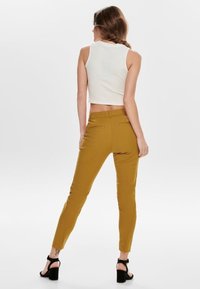 Mosterdgele stretchbroek met een aansluitend design, twee achterzakken en een gladde textuur, gecombineerd met een witte mouwloze top.