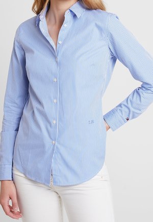 Kvinde iført en tætsiddende blå og hvid skjorte med lodrette striber, knapper og lange ærmer, samt hvide bukser, poserer med den ene hånd på hoften.