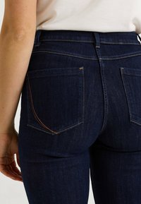 Vaqueros azul oscuro con una textura suave, que cuentan con dos bolsillos traseros y costuras de color naranja contrastante a lo largo de las costuras.