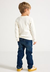 Maglietta bianca a maniche lunghe a coste abbinata a jeans blu scuro e scarpe da ginnastica gialle, vista da dietro, che mostra un design di abbigliamento casual.