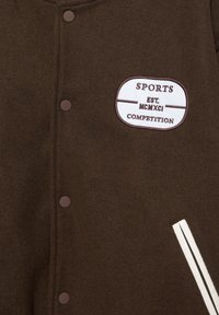 Hnědý kabát s kulatými knoflíky, bílou nášivkou s nápisem „SPORTS EST. MCMXCI COMPETITION“ a kapsou se světlým proužkem na pravé straně.