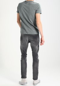 Man som bär en grå t-shirt, urblekta svarta jeans och vita sneakers, står vänd bort mot en enfärgad ljus bakgrund.