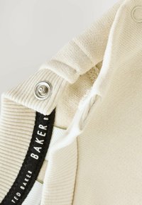 Beige kötött szövet bordázott textúrával, fémből készült patentgombbal és egy fekete címkével, amelyen fehér betűkkel az olvasható, hogy "TED BAKER."