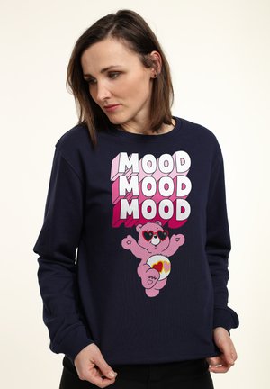 Námořnická modrá mikina s velkým grafickým designem růžového medvěda, který nosí sluneční brýle ve tvaru srdce, a nápisem "MOOD" psaným tučnými, pestrými písmeny.