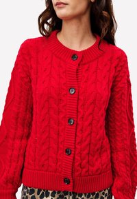 Cardigan rouge en tricot avec un motif en câble, présentant un col rond et cinq boutons noirs à l'avant. Poignets côtelés et ourlet inférieur.