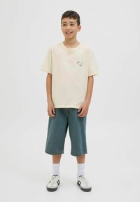 Helles beiges T-Shirt mit Brustdruck, kombiniert mit lockeren Denim-Shorts, weißen Socken und schwarz-weißen Sneakers. Einfaches Design und lässige Passform.