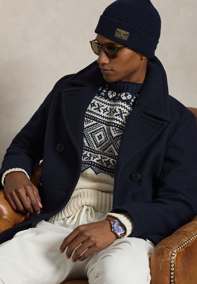 Μπλε ναυτική μάλλινη παλτό πάνω από κρεμ πλεκτό πουλόβερ με γεωμετρικό σχέδιο, συνδυασμένο με ανοιχτόχρωμο παντελόνι, μπλε σκούφο και γυαλιά ηλίου.