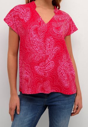 Vrouw draagt een rood topje met korte mouwen met roze paisley patronen en blauwe jeans, staand tegen een effen lichte achtergrond.