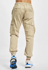 Persoon die beige cargobroek met elastische boorden en witte sneakers met blauwe en zwarte zolen draagt, lopend weg op een witte achtergrond.